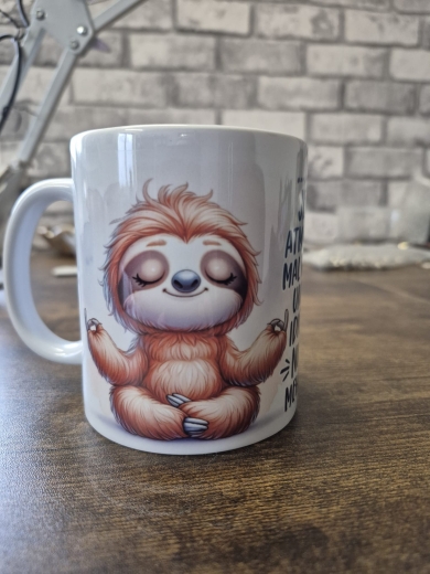 Zwei weiße Keramiktassen stehen auf einer hellen Holzoberfläche in einer ruhigen, dekorativen Umgebung. Beide Tassen zeigen ein meditierendes Faultier‑Comicmotiv mit geschlossenen Augen und entspanntem Gesichtsausdruck. Auf der linken Tasse befindet sich