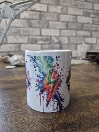 Eine weiße Keramiktasse steht auf einer Holzoberfläche vor einer unscharfen Wand mit Backsteinmuster. Auf der Tasse befindet sich ein auffälliges, farbenfrohes Lippen‑Motiv mit kräftigen Farbverläufen und Farbklecks‑Effekten in Blau, Rot, Grün und Lila. D