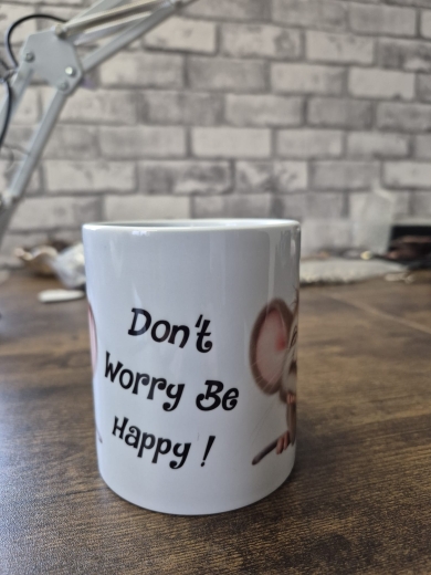Eine weiße Keramiktasse steht auf einer Holzoberfläche vor einer Wand mit Backsteinmuster. Auf der Vorderseite der Tasse befindet sich der gut lesbare Schriftzug „Don’t Worry Be Happy!“, daneben ist ein teilweise sichtbares Comic‑Tiermotiv mit großen Ohre