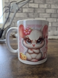 Eine weiße Keramiktasse steht auf einer Holzoberfläche vor einer unscharfen Wand mit Backsteinmuster. Auf der Vorderseite befindet sich der auffällige pinke Schriftzug „MINI MOTZ MAUS“, umgeben von kleinen Herzmotiven. Links und rechts daneben sind zwei n