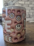 Eine weiße Keramiktasse steht auf einer Holzoberfläche vor einer unscharfen Backsteinwand. Die Tasse zeigt ein farbenfrohes Design mit zwei illustrierten Kaffeebechern, dekorativen Blumenmotiven und dem gut lesbaren Spruch „ALL YOU NEED IS LOVE AND A GOOD