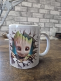 Eine weiße Keramiktasse steht auf einer Holzoberfläche vor einem grauen Hintergrund. Auf der Tasse befindet sich ein farbenfrohes Baby‑Groot‑Motiv, das den Eindruck vermittelt, als würde die Figur durch eine aufgebrochene Wand brechen. Die kräftigen Farbe
