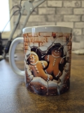Eine weiße Keramiktasse steht auf einer Holzoberfläche vor einer unscharfen Wand mit Backsteinmuster. Auf der Tasse befindet sich ein farbenfrohes Lebkuchen‑Motiv mit zwei fröhlichen Figuren, die scheinbar durch eine aufgebrochene Ziegelwand brechen. Die