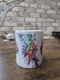 Eine weiße Keramiktasse steht auf einer Holzoberfläche vor einer unscharfen Wand mit Backsteinmuster. Auf der Tasse befindet sich ein auffälliges, farbenfrohes Lippen‑Motiv mit kräftigen Farbverläufen und Farbklecks‑Effekten in Blau, Rot, Grün und Lila. D