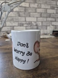 Eine weiße Keramiktasse steht auf einer Holzoberfläche vor einer Wand mit Backsteinmuster. Auf der Vorderseite der Tasse befindet sich der gut lesbare Schriftzug „Don’t Worry Be Happy!“, daneben ist ein teilweise sichtbares Comic‑Tiermotiv mit großen Ohre